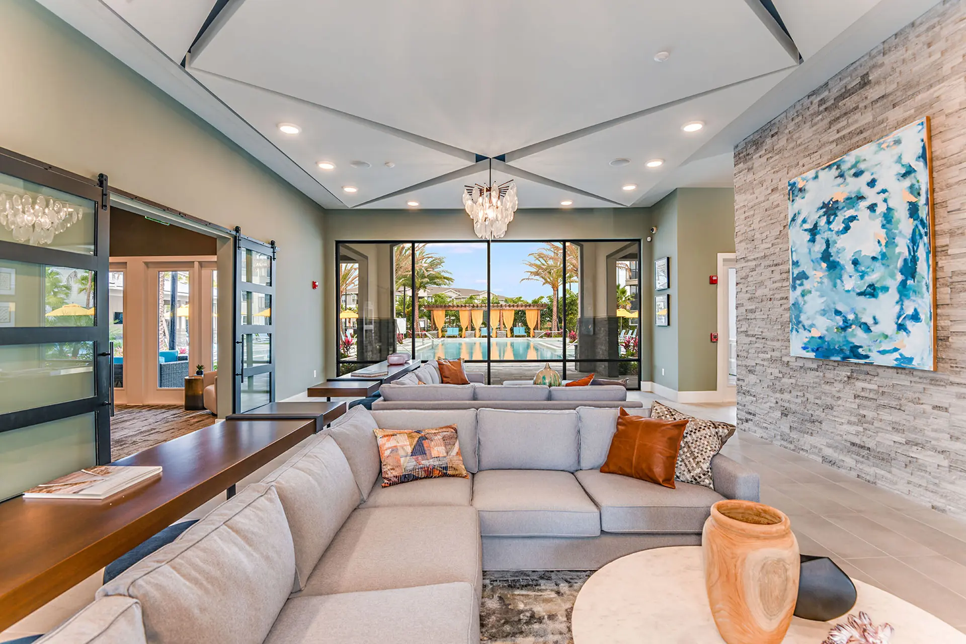 Indoor lounge area with chandelier, two large gray, L-shaped couches, coffee tables, and a ledge-stone accent wall.