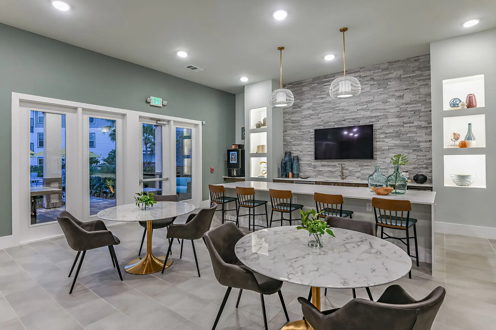 luxury apartment clubouse with large island bar, large TV on a ledge stone wall, two marble dining tables, and a view of the swimming pool area.