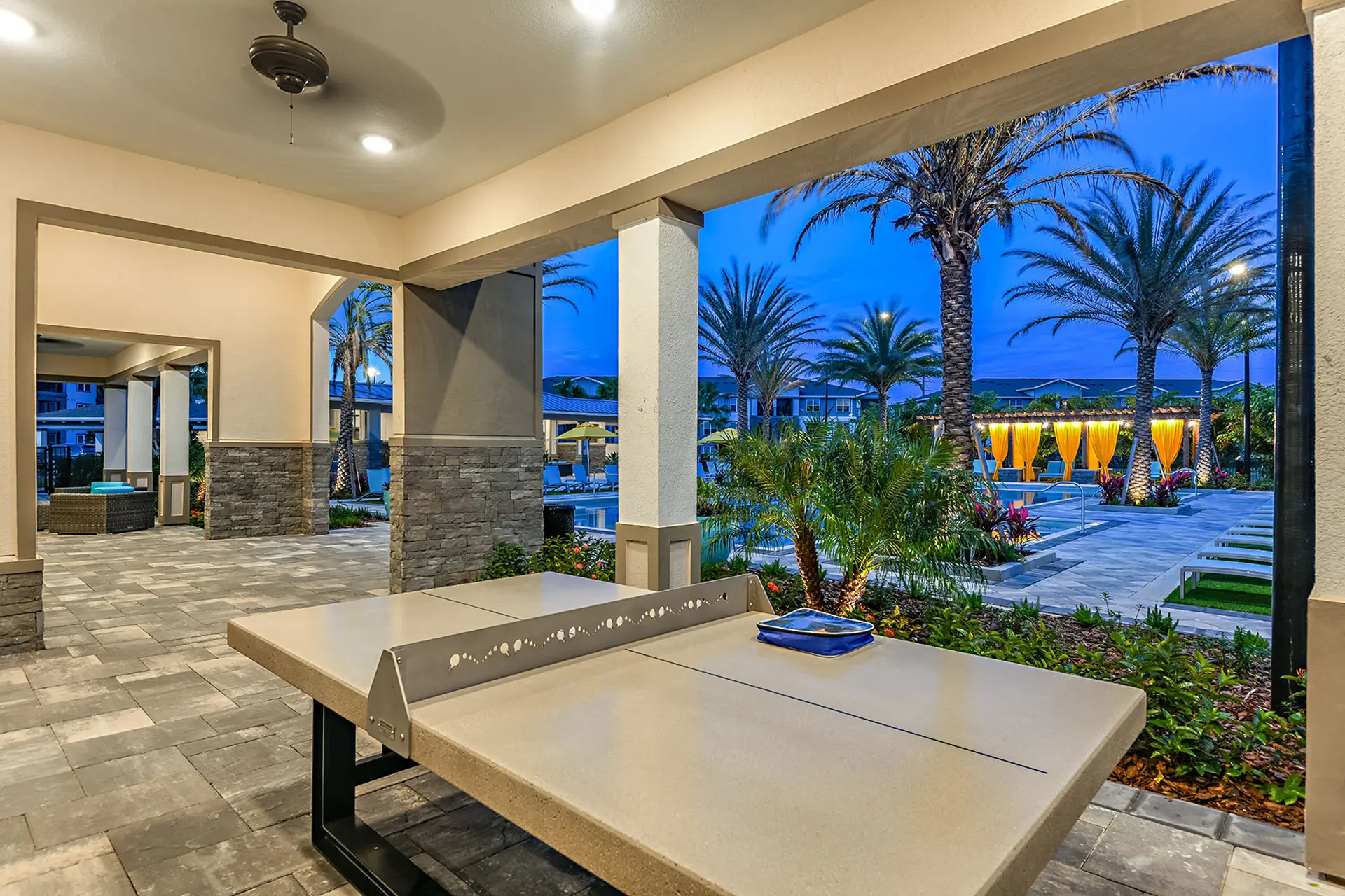 outdoor cement pin-pong table next to a large swimming pool surrounded by palm trees.