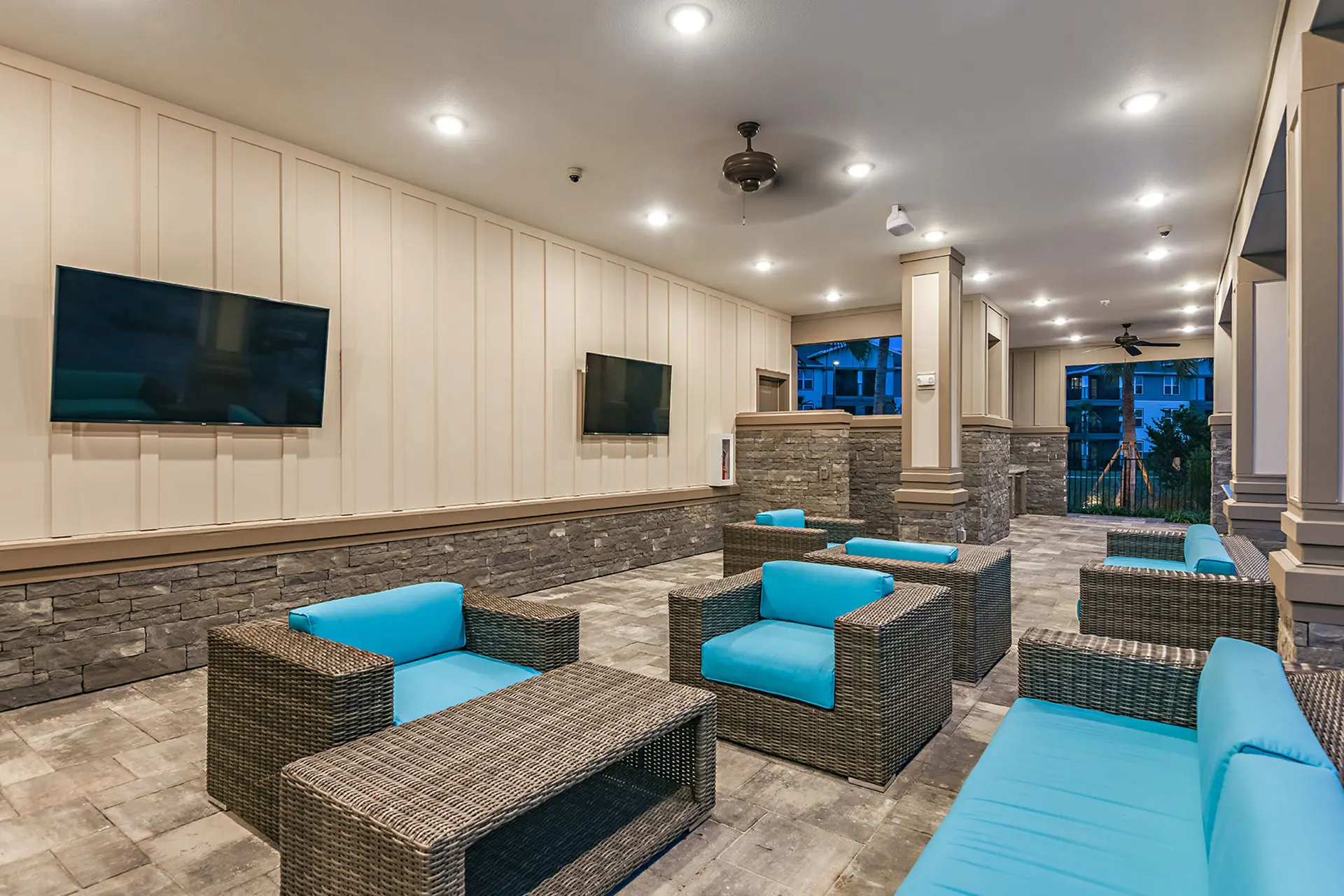 outdoor covered stone patio with blue wicker couches, two large TVs, recessed overhead lighting, and ceiling fans.