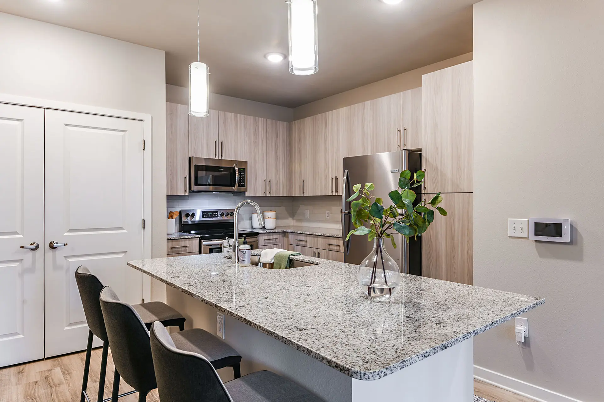 apartment kitchen with light upgraded cabinets with red undertones, stainless appliances, and an island with sink, granite countertop, and three bar chairs.