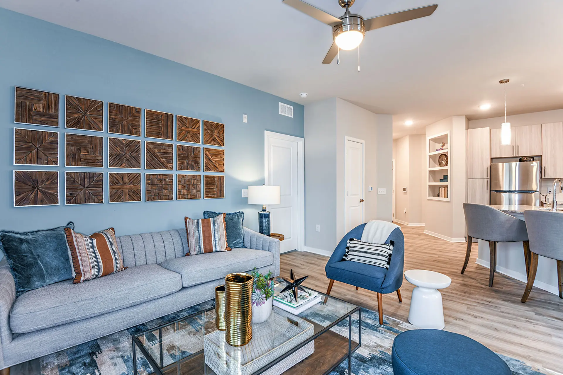 apartment living room with wood style flooring, a light-blue accent wall, blue chairs, a glass coffee table, and a light-blue sofa.