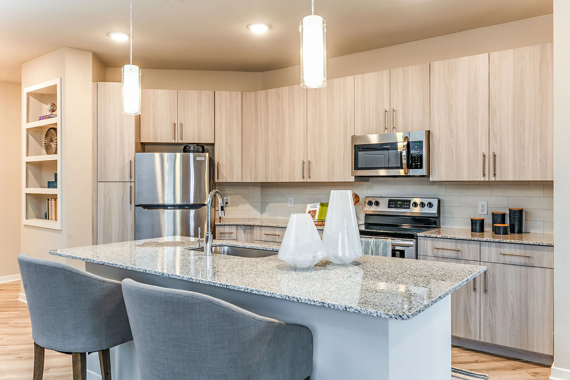 apartment kitchen with large island with granite countertop, two bar chairs, upgraded cabinets, pendant lighting, and stainless appliances.