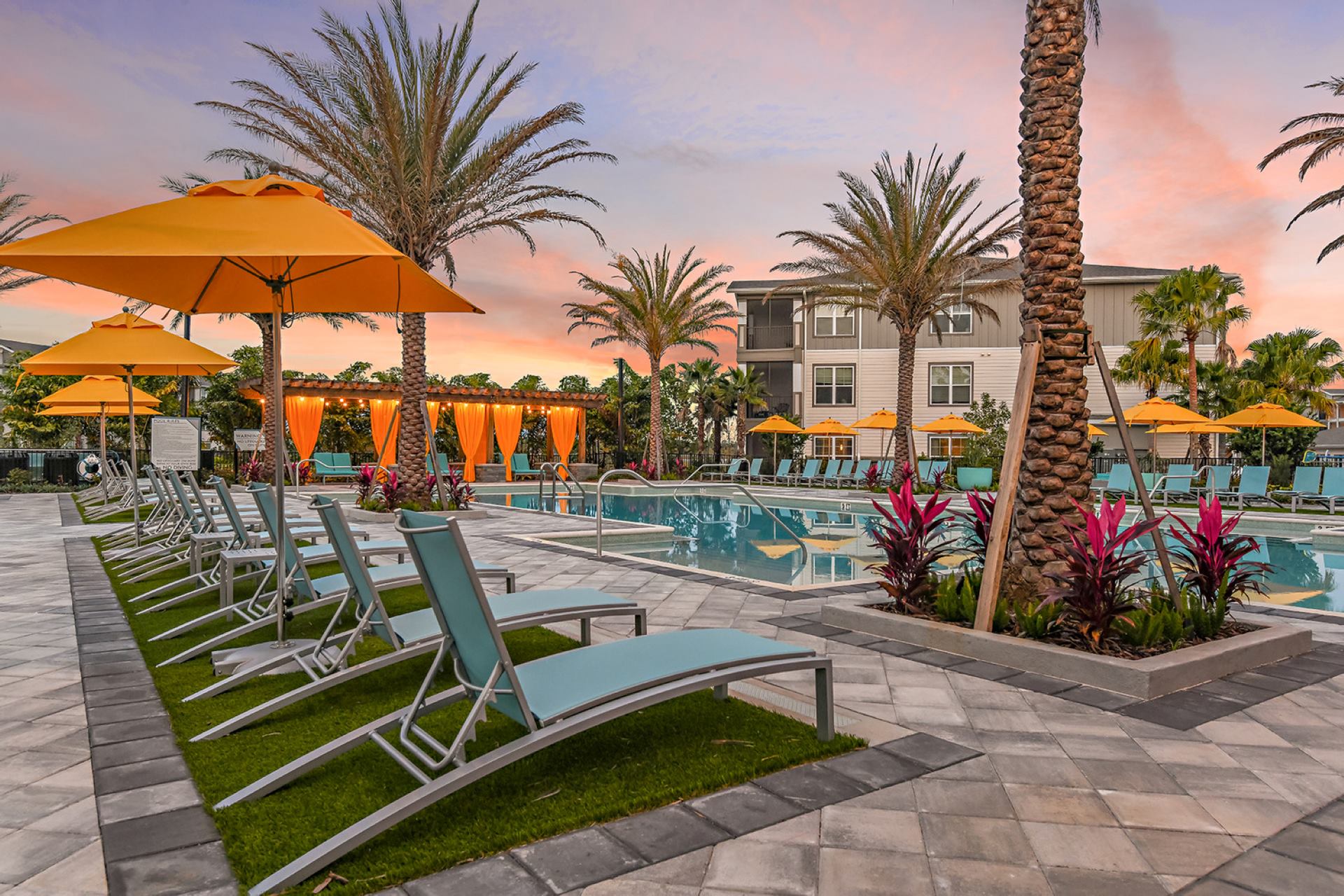 Large pool and sundeck, blue lounge chairs, orange umbrellas, native Florida landscaping, palm trees